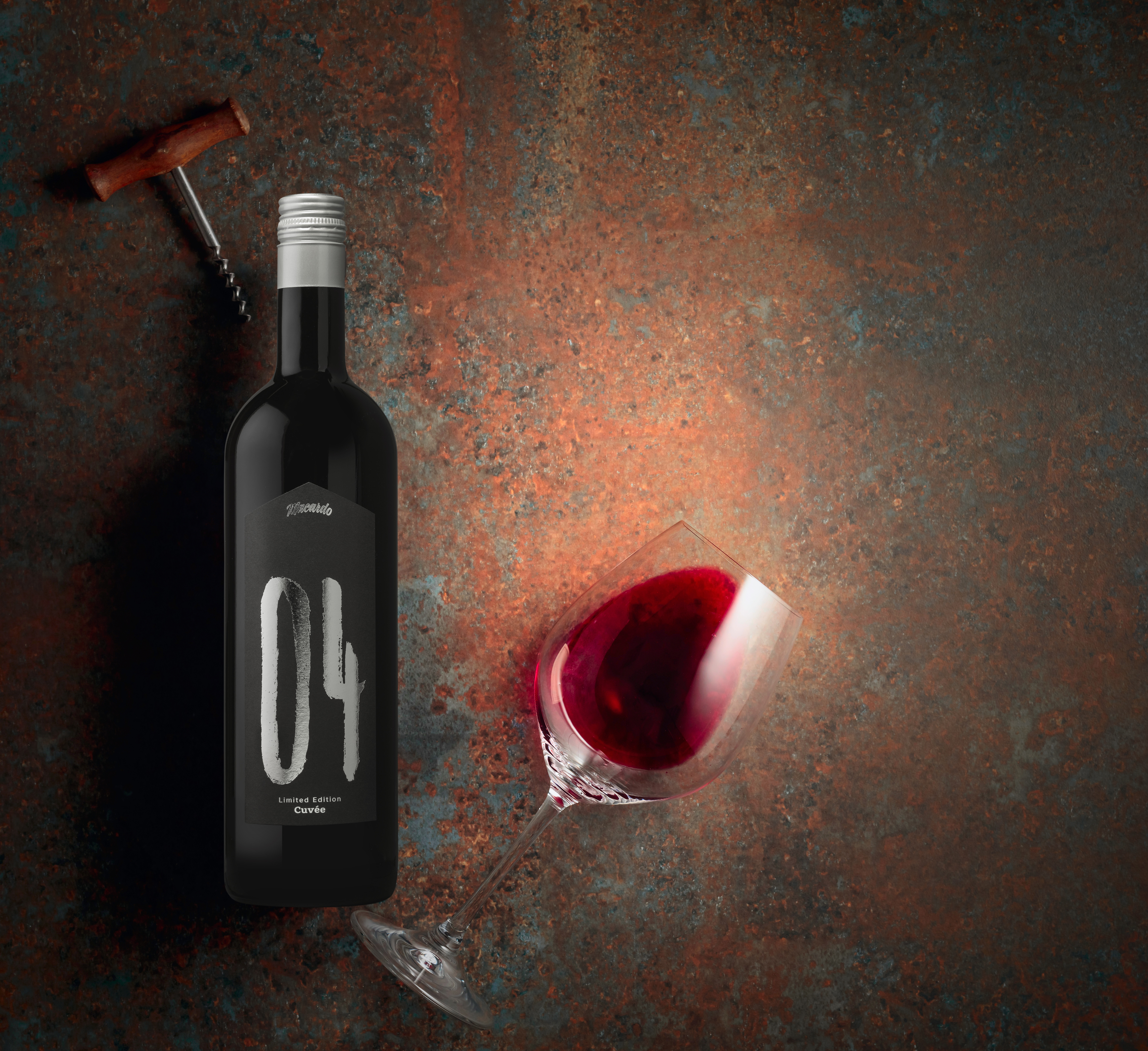 Eine Flasche "04" Cuvée Rotwein mit einem Korkenzieher und einem Glas Rotwein auf einem rustikalen, mehrfarbigen Untergrund. Auf dem Etikett der Flasche steht gut sichtbar die Nummer "04".